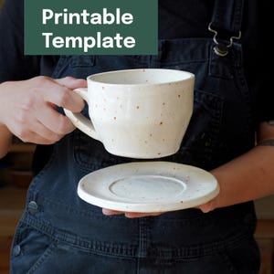 Può includere: Una tazza in ceramica bianca macchiata con un piattino abbinato. La tazza ha un manico grande e un'apertura ampia. Il piattino è rotondo con un bordo leggermente rialzato. Il testo "Printable Template" è in un rettangolo verde sopra la tazza.