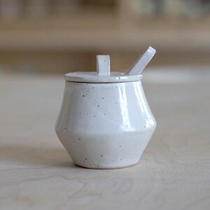 May include: A white ceramic sugar bowl with a lid and a small spoon. The bowl has a distinctive, geometric shape and a speckled texture. The spoon rests on the side of the bowl. The sugar bowl is on a light-coloured wooden surface.