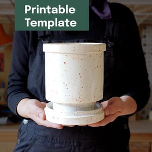 Op de afbeelding: Een gespikkelde, crèmekleurige keramische container met een deksel en een basis. De container heeft een cilindrische vorm met een iets bredere basis en deksel. De tekst "Printable Template" staat in een groene rechthoek linksboven.