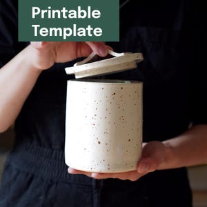 May include: A speckled, off-white ceramic jar with a matching lid is held open. The jar is cylindrical with a slightly textured surface and small brown speckles. The text "Printable Template" is in a green rectangle in the upper left.