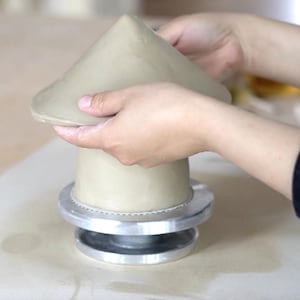 Pu&ograve; includere: Scultura in argilla beige chiaro in fase di formazione. La base cilindrica poggia su una base in metallo argentato. La parte superiore conica &egrave; modellata a mano, mostrando l'artigianato. Probabile progetto di ceramica.