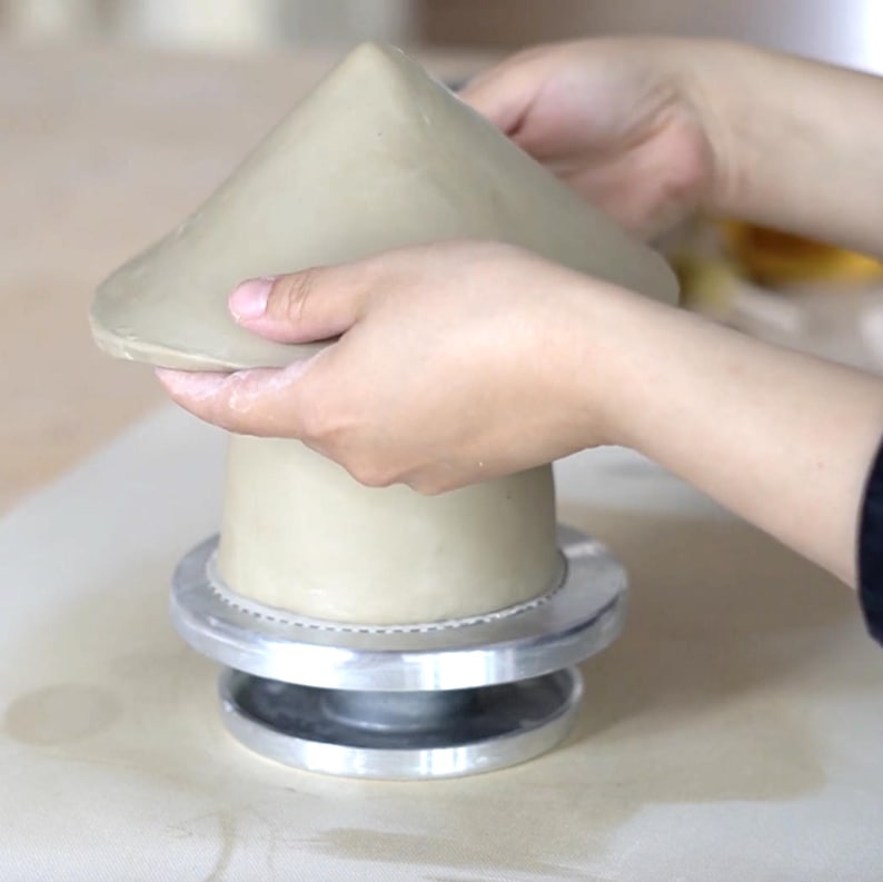 Pu&ograve; includere: Scultura in argilla beige chiaro in fase di formazione. La base cilindrica poggia su una base in metallo argentato. La parte superiore conica &egrave; modellata a mano, mostrando l'artigianato. Probabile progetto di ceramica.