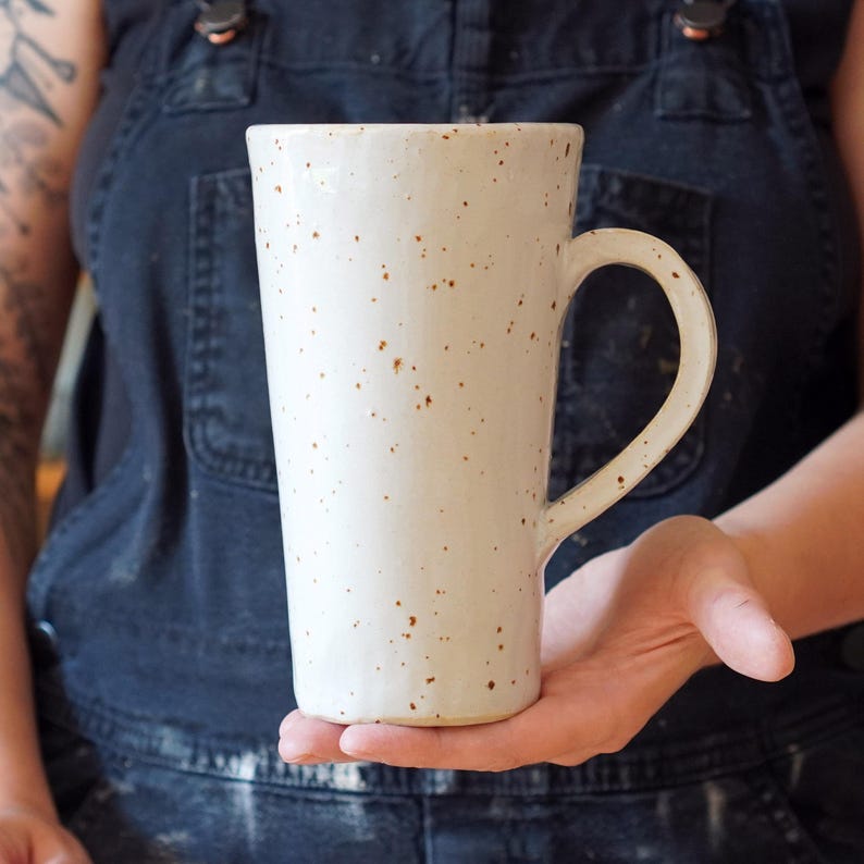 K&ouml;nnte beinhalten: Eine wei&szlig;e Keramiktasse mit braunen Flecken und einer hohen, zylindrischen Form. Die Tasse hat einen gro&szlig;en Henkel.