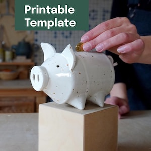 Può includere: Un salvadanaio in ceramica bianca con macchie marroni, a forma di maiale, è posto su un blocco di legno chiaro. Il salvadanaio ha una fessura sulla parte superiore per inserire le monete. Il testo "Printable Template" è in un rettangolo verde in alto a sinistra.