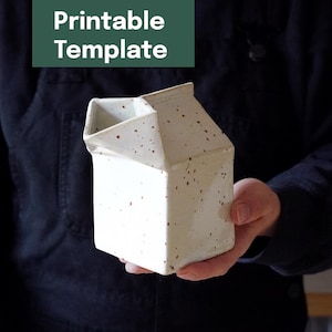 May include: A ceramic milk carton shaped pitcher with a white glaze and brown speckles. The top features a folded spout design. The text Printable Template is displayed in white on a dark green rectangular background at the top of the image.