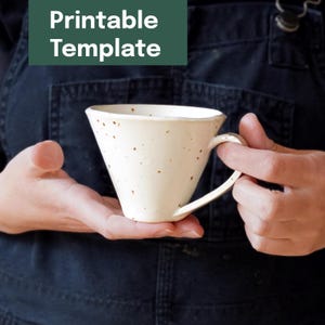 Puede incluir: Una taza de cerámica color crema con un diseño angular único. La taza tiene un asa y está salpicada de manchas marrones. El texto "Printable Template" está en un rectángulo verde encima de la taza.