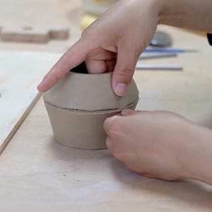 May include: A light brown clay pot in the process of being shaped by hand. The pot has a wide base, a narrower middle section, and a flared top. Hands are smoothing the clay, creating a handmade ceramic piece.