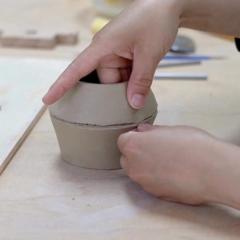 May include: A light brown clay pot in the process of being shaped by hand. The pot has a wide base, a narrower middle section, and a flared top. Hands are smoothing the clay, creating a handmade ceramic piece.