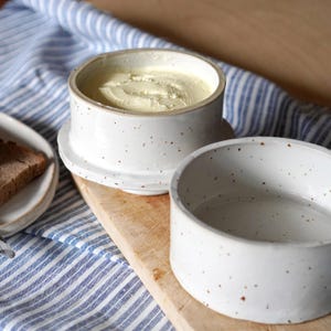 Op de afbeelding: Witte keramische botervloot met gespikkeld glazuur. De vloot heeft een deksel en een aparte container om boter in te bewaren.
