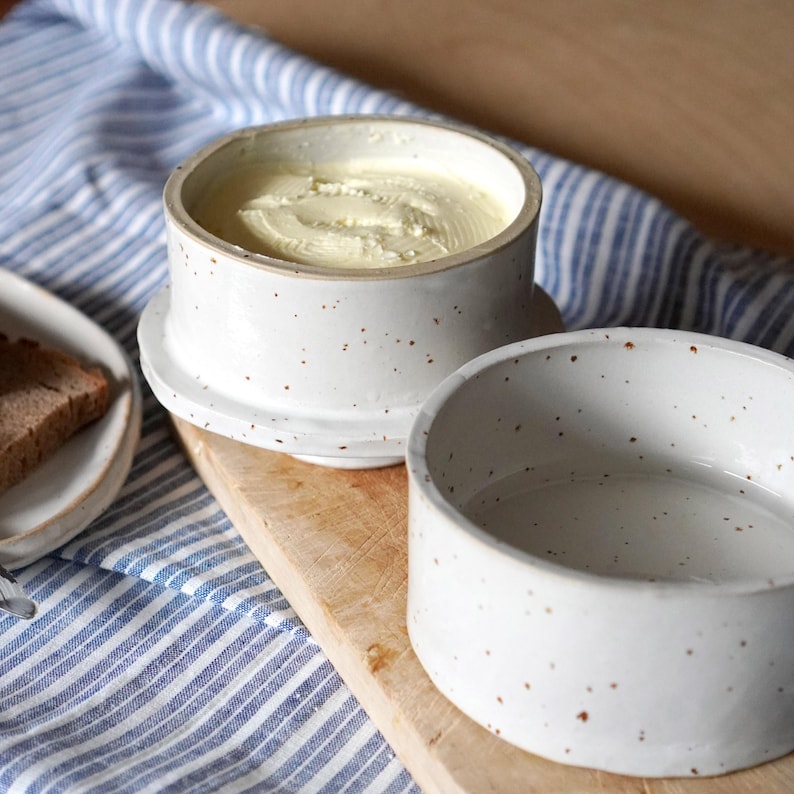 Op de afbeelding: Witte keramische botervloot met gespikkeld glazuur. De vloot heeft een deksel en een aparte container om boter in te bewaren.