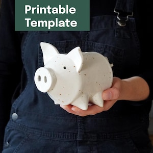 Könnte beinhalten: Ein weißes Keramik-Sparschwein mit braunen Sprenkeln wird in einer Hand gehalten. Das Sparschwein hat einen runden Körper, vier kleine Beine und eine Schnauze mit zwei Löchern. Der Text "Printable Template" befindet sich in einem grünen Rechteck im Hintergrund.