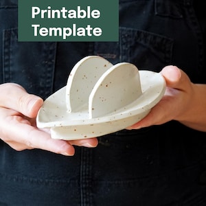 Puede incluir: Un organizador de escritorio de cerámica blanca moteada con dos divisores curvos y una base redondeada. El texto "Printable Template" está en un rectángulo verde en la esquina superior izquierda. El organizador está en las manos de una persona.
