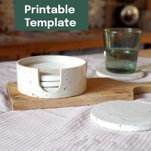 Pode incluir: Um conjunto de quatro bases de cerâmica branca com salpicos castanhos, guardadas num suporte a condizer. As bases e o suporte estão sobre uma tábua de cortar de madeira. O texto na imagem diz "Printable Template."