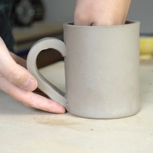 May include: A grey ceramic mug with a handle being shaped by hand. The mug is sitting on a light brown surface.