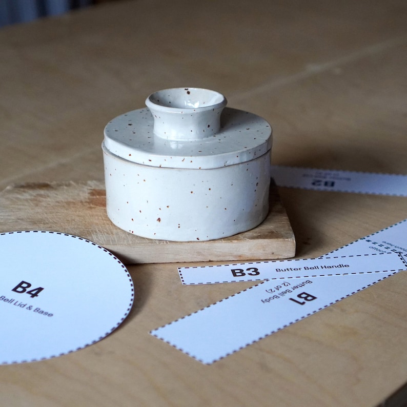 Op de afbeelding: Witte keramische botervloot met een gespikkelde glazuur en een bruine handgreep. De botervloot is gelabeld met "B3 Butter Bell Handle" en "B1 Butter Bell Body".