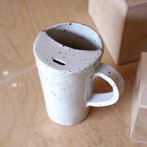 K&ouml;nnte beinhalten: Eine wei&szlig;e Keramiktasse mit braunen Flecken und einem einzigartigen Henkel. Die Tasse hat einen Ausguss an der Seite und einen leicht unebenen Rand.