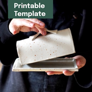 Könnte beinhalten: Eine gesprenkelte Keramik-Butterdose mit Deckel, gehalten von einer Person. Die Dose ist rechteckig mit abgerundeten Kanten und einem Griff am Deckel. Der Text "Printable Template" befindet sich in einem grünen Rechteck oben.