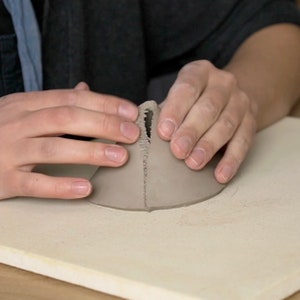 May include: A person's hands are holding a grey clay cone with a small hole at the top. The cone is sitting on a white surface.