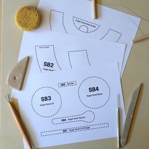 May include: White paper templates for a sugar bowl, including the base, lid, handle, and spoon. The templates are labelled with dotted lines and text. A yellow sponge and pottery tools are also present.