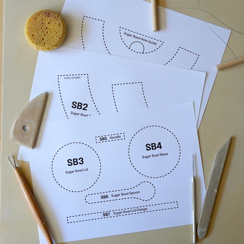 May include: White paper templates for a sugar bowl, including the base, lid, handle, and spoon. The templates are labelled with dotted lines and text. A yellow sponge and pottery tools are also present.
