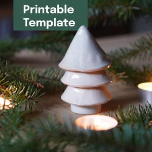 Könnte beinhalten: Ein weißer Weihnachtsbaum aus Keramik mit einem geschichteten Design. Der Baum ist von Grün und brennenden Kerzen umgeben. Der Text "Printable Template" befindet sich in einem grünen Rechteck über dem Baum.