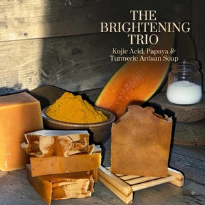 May include: The Brightening Trio Kojic Acid, Papaya & Turmeric Artisan Soap set includes several rectangular bars of orange-toned soap, a bowl of yellow turmeric powder, a slice of orange papaya, and a small glass jar containing white powder.