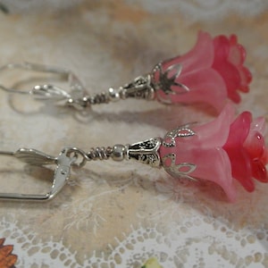 May include: A pair of silver earrings with pink flower-shaped charms. The flowers have a layered design and are translucent.