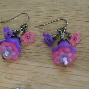 May include: A pair of dangle earrings with purple and pink flower-shaped beads. The earrings are made of metal and have a silver finish.