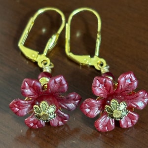 May include: A pair of dangle earrings featuring burgundy flower-shaped pendants with gold-colored accents. The earrings have gold-colored lever-back closures and small red beads. The flower petals have a textured appearance.