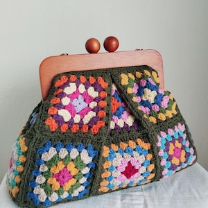 May include: A crochet handbag featuring a multi-coloured granny square pattern in shades of pink, blue, yellow, and orange, framed by a dark olive green border. The bag has a wooden frame with two round wooden beads as a clasp closure mechanism.