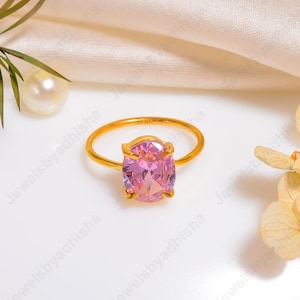 Puede incluir: Un anillo de oro con una gran gema rosa de talla ovalada. El anillo tiene una banda fina y sencilla, y la gema est&aacute; engastada en un engaste de cuatro puntas. El anillo se exhibe sobre una superficie blanca con perlas y detalles florales.