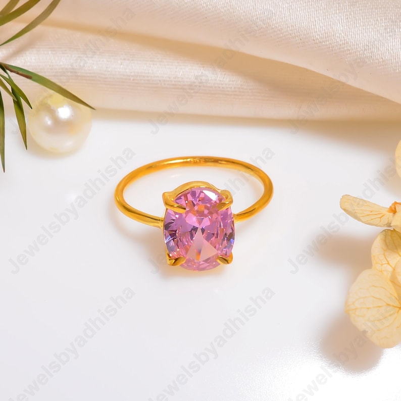 Puede incluir: Un anillo de oro con una gran gema rosa de talla ovalada. El anillo tiene una banda fina y sencilla, y la gema est&aacute; engastada en un engaste de cuatro puntas. El anillo se exhibe sobre una superficie blanca con perlas y detalles florales.