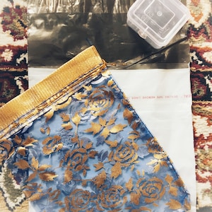 Puede incluir: Una tela azul con un estampado floral dorado, doblada y colocada sobre un sobre blanco. La tela tiene un borde dorado. En la imagen tambi&eacute;n hay un peque&ntilde;o recipiente de pl&aacute;stico transparente y un sobre de pl&aacute;stico negro.