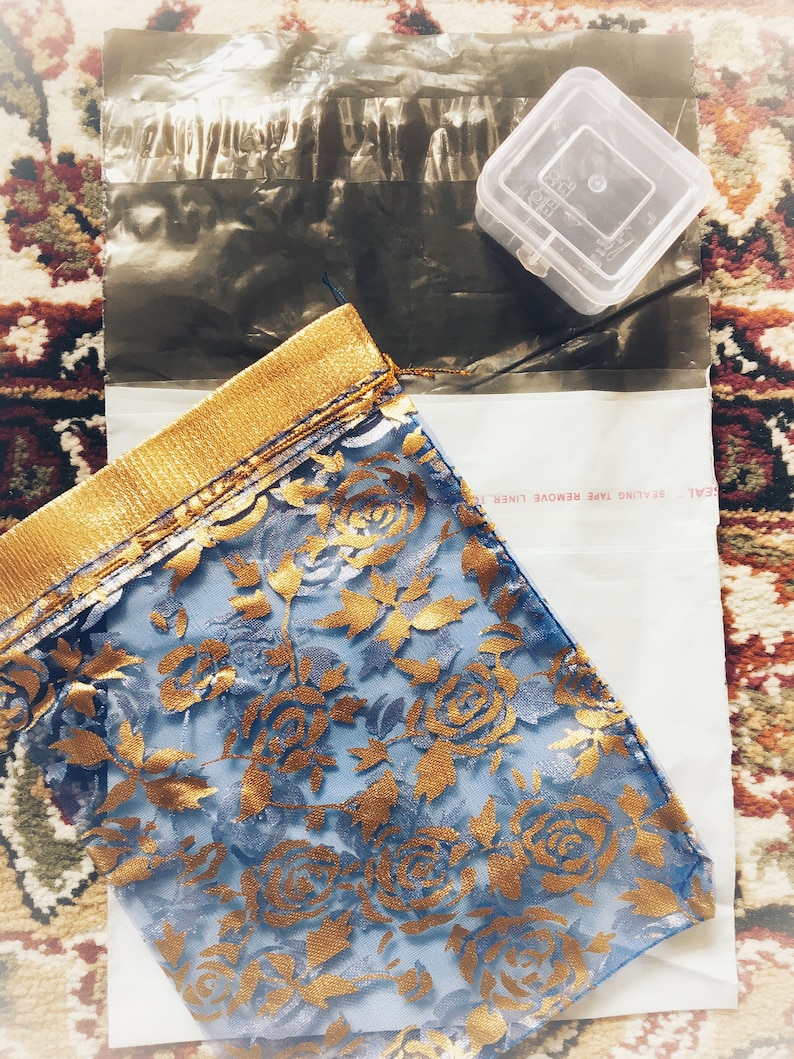 Puede incluir: Una tela azul con un estampado floral dorado, doblada y colocada sobre un sobre blanco. La tela tiene un borde dorado. En la imagen tambi&eacute;n hay un peque&ntilde;o recipiente de pl&aacute;stico transparente y un sobre de pl&aacute;stico negro.