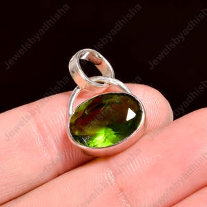 May include: A silver pendant featuring an oval, faceted, olive-green gemstone. The gemstone is set in a silver bezel with a loop for a chain. The pendant is held in a hand against a dark background. The jewelry is from Jewelsbyadhisha.