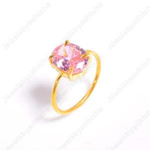 Puede incluir: Un anillo dorado con una gran gema rosa de corte ovalado. La banda y el engaste del anillo son dorados, creando un contraste con la piedra rosa claro. El anillo est&aacute; sobre un fondo blanco.