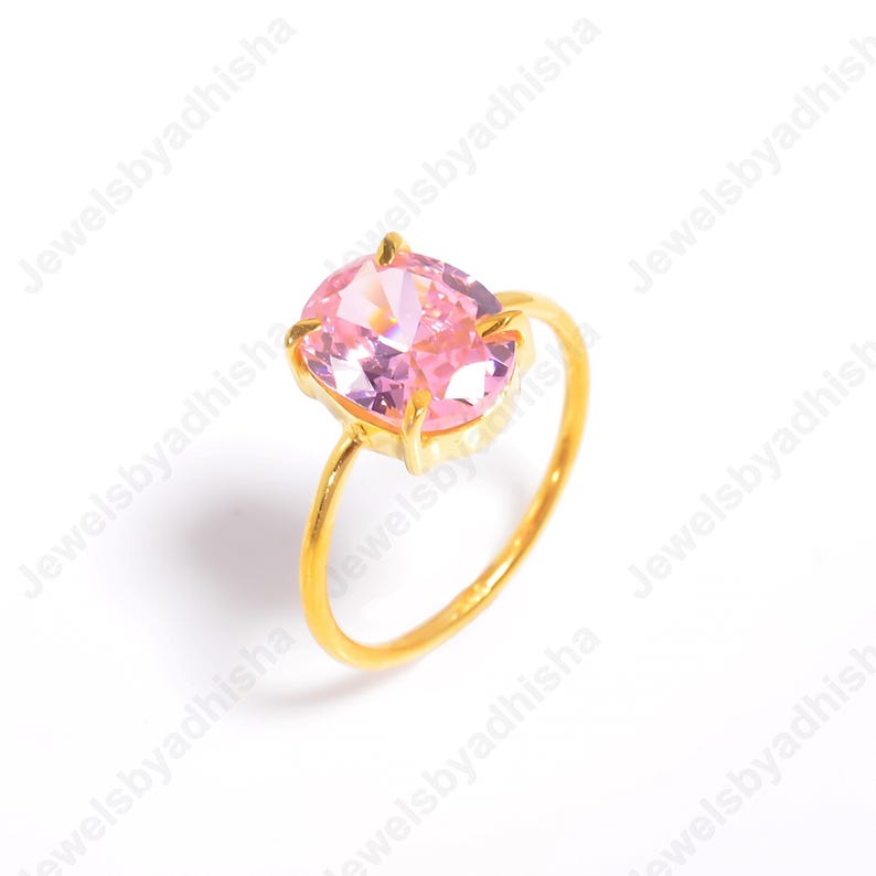 Puede incluir: Un anillo dorado con una gran gema rosa de corte ovalado. La banda y el engaste del anillo son dorados, creando un contraste con la piedra rosa claro. El anillo est&aacute; sobre un fondo blanco.