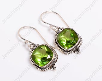 May include: A pair of silver drop earrings featuring square-cut, light green gemstones. The gemstones are set in a decorative, textured silver frame with a hook closure. The earrings are displayed on a white surface.