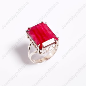 May include: A silver ring featuring a large, rectangular, vibrant red gemstone. The gemstone is set in a detailed, openwork silver frame, with a simple band. The ring is displayed on a white background.