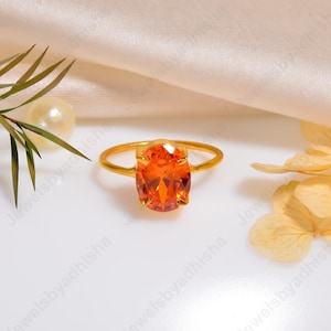 Könnte beinhalten: Ein goldener Ring mit einem ovalen, orangefarbenen Edelstein. Band und Fassung des Rings sind goldfarben, und der Edelstein ist der Blickfang. Der Ring wird auf einer weißen Oberfläche mit dekorativen Elementen präsentiert.