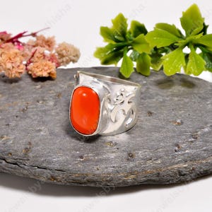 May include: A silver ring featuring a smooth, oval-shaped orange coral gemstone set in a polished metal band. The side of the band displays an embossed Om symbol, providing a detailed contrast to the vibrant, solid-colored stone.