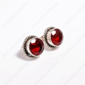 May include: A silver ring with two large, round, deep red gemstones. The gemstones are set in a bezel setting and surrounded by a twisted rope design. The ring is shown against a white backdrop.