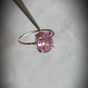 Puede incluir: Un anillo de plata con una gran gema rosa de talla ovalada. El anillo es sujetado por unas pinzas, mostrando las facetas pulidas de la piedra. El fondo es de un color suave y neutro, resaltando la joya.