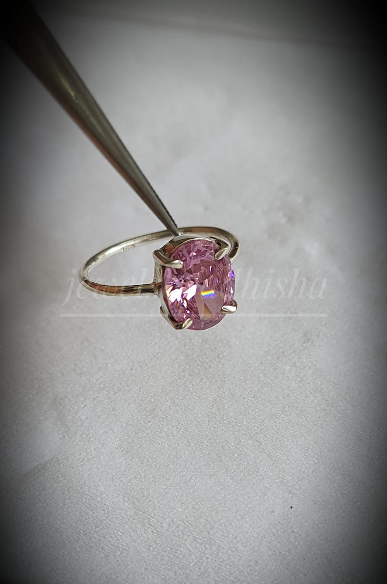 Puede incluir: Un anillo de plata con una gran gema rosa de talla ovalada. El anillo es sujetado por unas pinzas, mostrando las facetas pulidas de la piedra. El fondo es de un color suave y neutro, resaltando la joya.