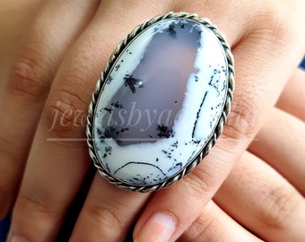 Può includere: Un anello d'argento con una grande pietra di agata bianca ovale con inclusiones dendritiche nere. L'anello ha un design a corda attorno al bordo della pietra.