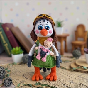 May include: A handmade crochet bird doll wearing a brown aviator hat and green dress. The bird has orange feet and a white body. It is holding a smaller doll. The background includes books and a small table.