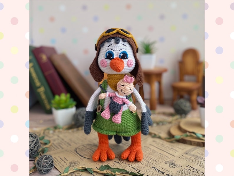 May include: A handmade crochet bird doll wearing a brown aviator hat and green dress. The bird has orange feet and a white body. It is holding a smaller doll. The background includes books and a small table.
