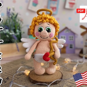 Puede incluir: Muñeco amigurumi de crochet con halo, alas y arco y flecha. El muñeco lleva un pañal y tiene un corazón rojo en su flecha. El muñeco está sentado sobre una base de madera.