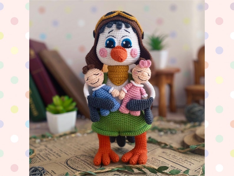 May include: A handmade crochet bird doll wearing a pilot's cap and holding two smaller dolls. The bird has a white face, blue eyes, and an orange beak and feet. It is wearing a green skirt and a yellow scarf.