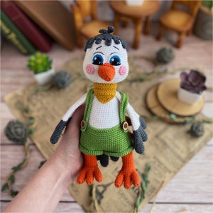 May include: A handmade crochet bird doll with a white body, black and gray wings, and orange feet. The bird wears green overalls with wooden buttons and a yellow neckerchief. The doll has blue eyes and an orange beak.